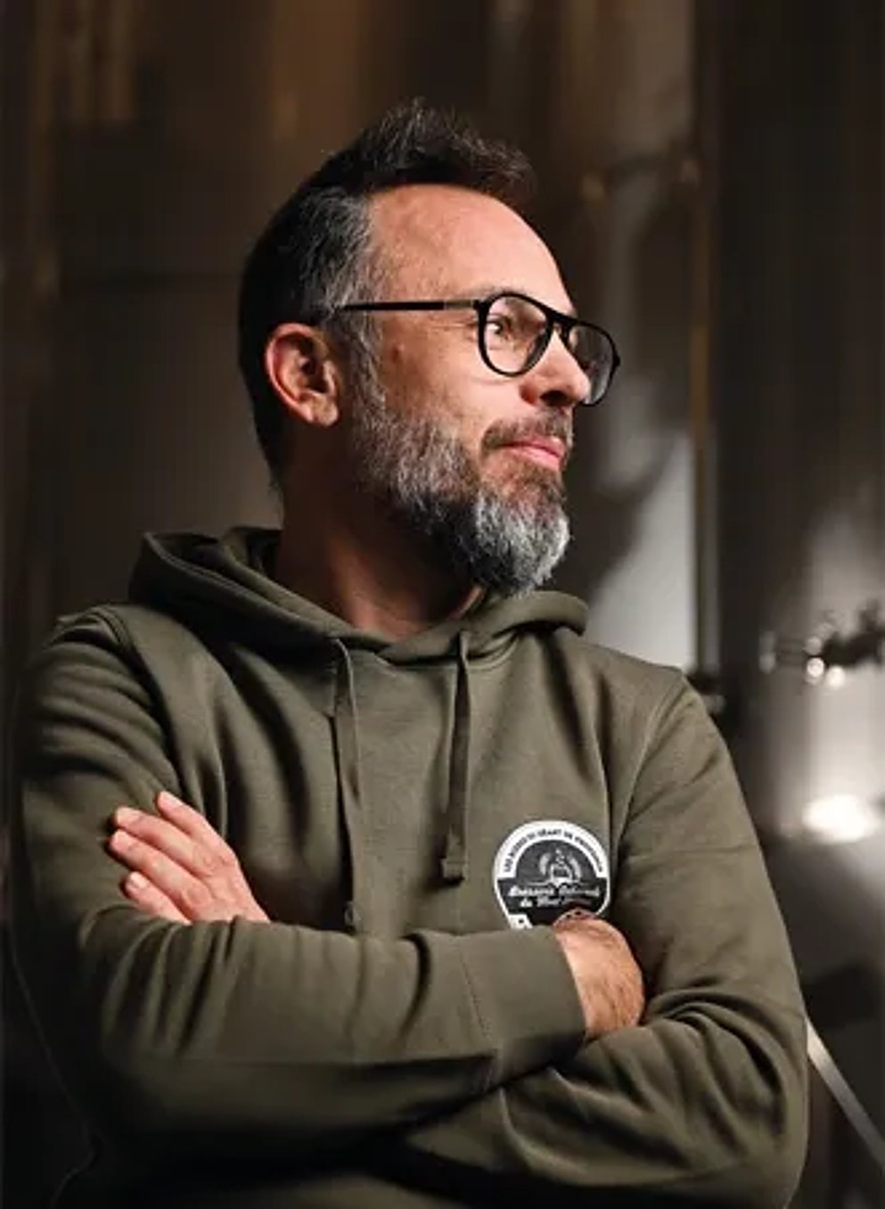 Un brasseur barbu à lunettes, en sweat kaki, devant les cuves